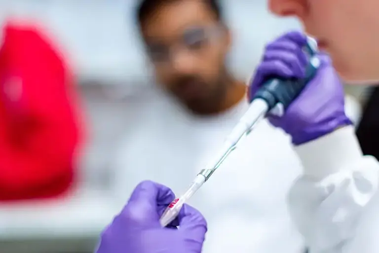 Close up of a scientist using a pipette in a lab, while a second scientist looks on in the background