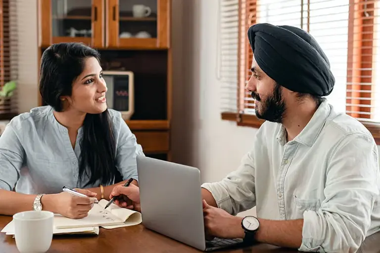 A Sikh couple talk to each other sitting at a dining room table, one with an open laptop point to lines in the other's open notebook 