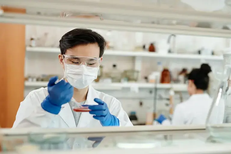 Two scientists in a lab, one the background and the one in the foreground adding material to a petri dish.
