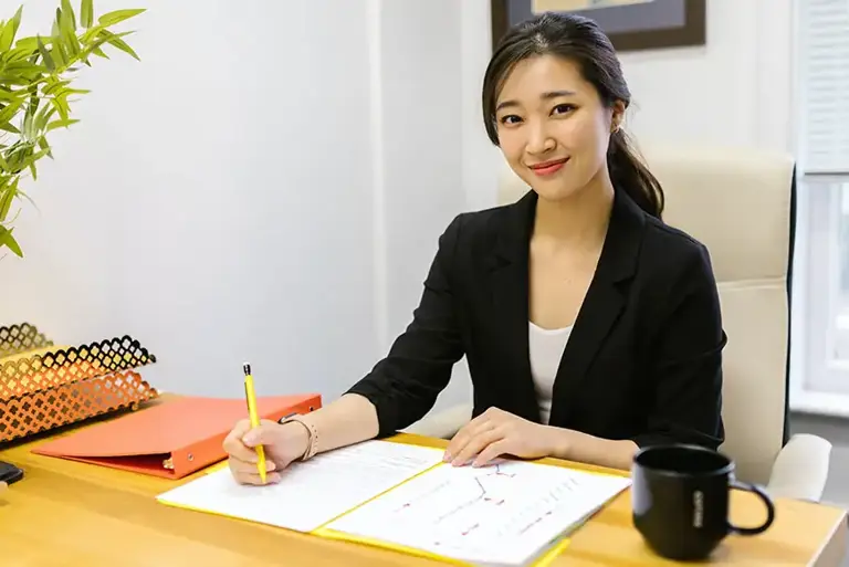 A smiling office worker sits a desk, writing in her notebook 