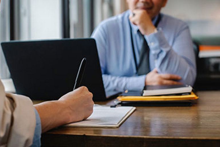 Man sitting at a desk with this laptop speaking to an interviewer who is taking notes