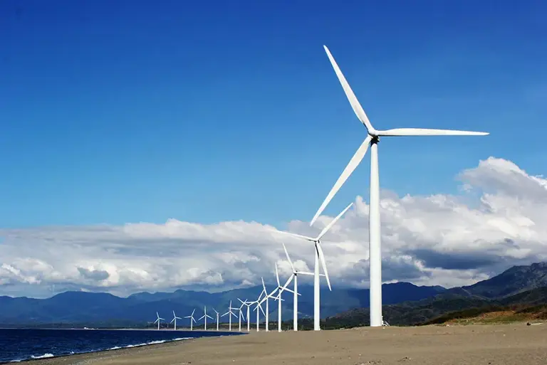 Series of wind turbines along a beach stretch into the distance lapped by a blue sea against bright blue sky.