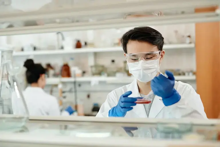 Two scientists in a lab, one the background and the one in the foreground adding material to a petri dish.