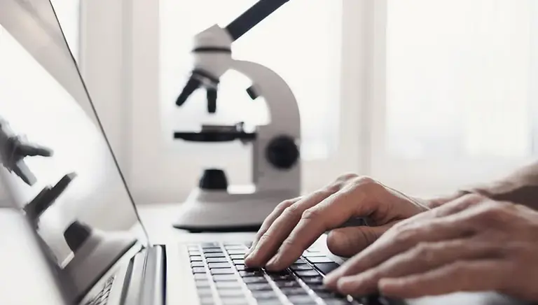 Close-up of hands typing on a laptop, with a microscope in the background, being reflected on the screen