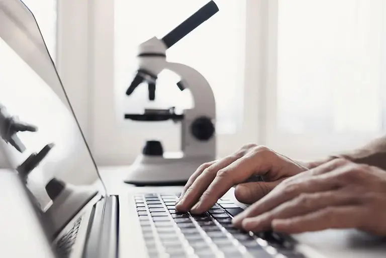 Close-up of hands typing on a laptop, with a microscope in the background, being reflected on the screen