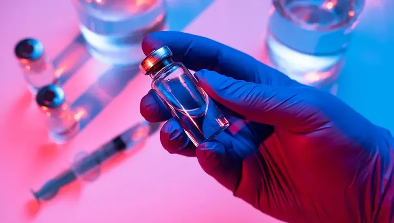 Close-up of a purple surgical gloved hand holding a vial filled with a clear liquid, above a table top with more vials 