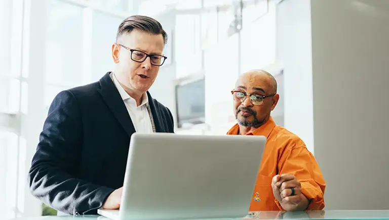 Two office workers at a standing desk are discussing a the issue they see on the laptop before them