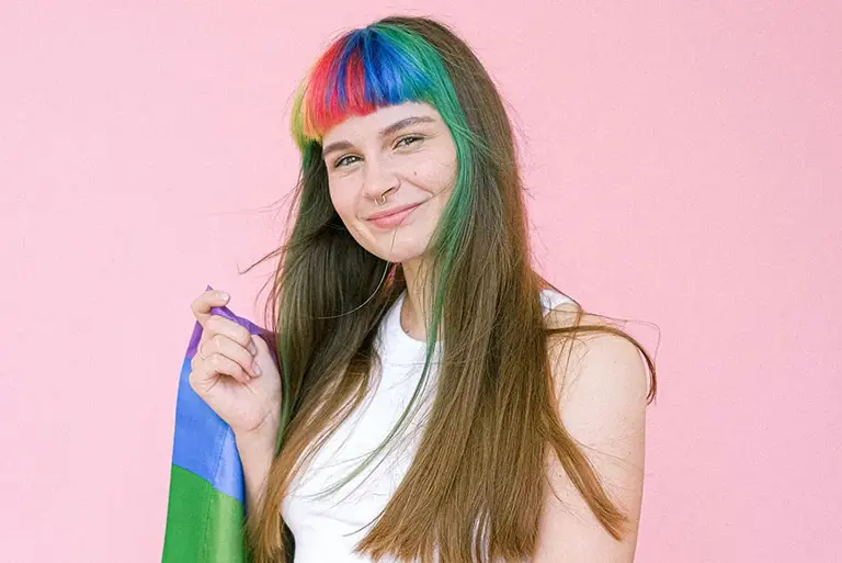 Young smiling woman with a rainbow coloured fringe is wrapped in a rainbow flag against a pink background.
