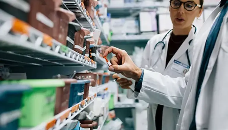 Two pharmacists face a full prescription medicines rack, while examining one particular medicine