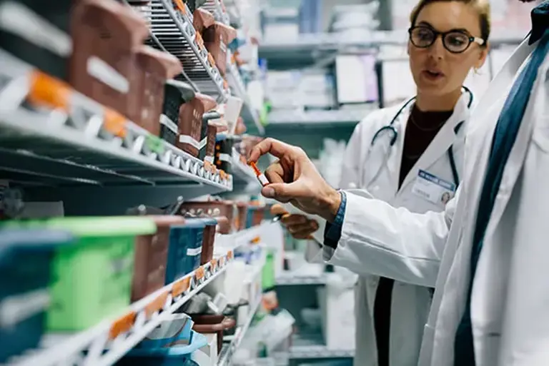 Two pharmacists face a full prescription medicines rack, while examining one particular medicine