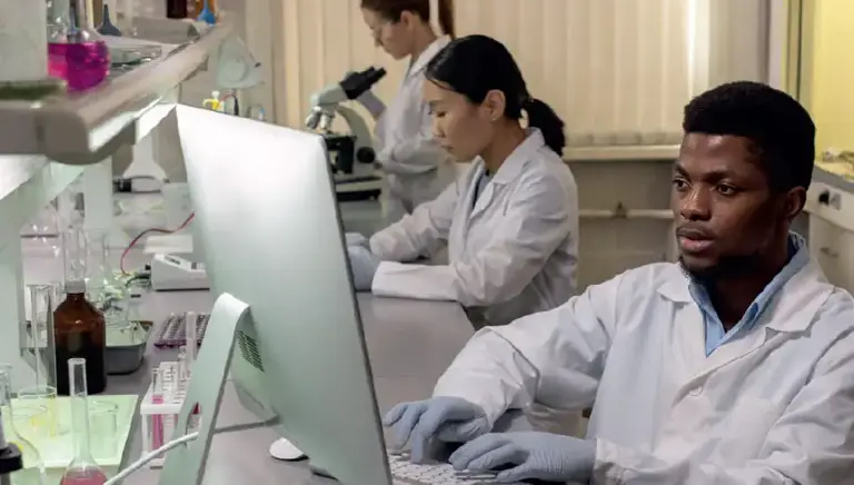Three scientists are working in a lab, one on a microscope, one with a pipette and the final one works on a laptop 