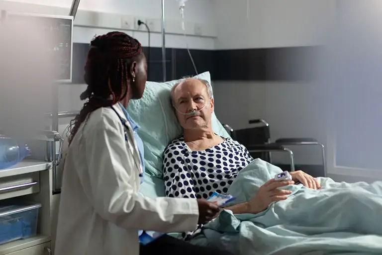 Doctor sits next to an older patient lying in a bed in a hospital setting