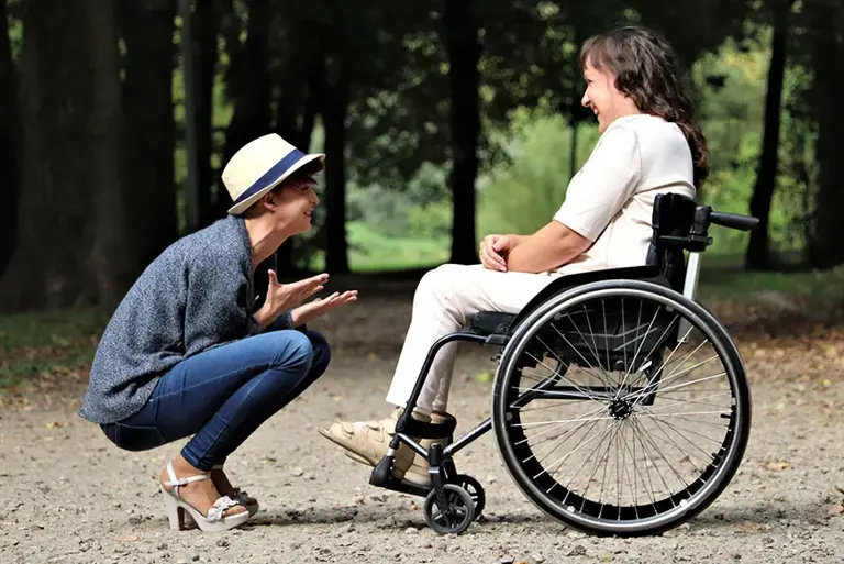A smiling woman sat in a wheelchair is smiling at young person, in women's high heels, who is crouching in front of her. 