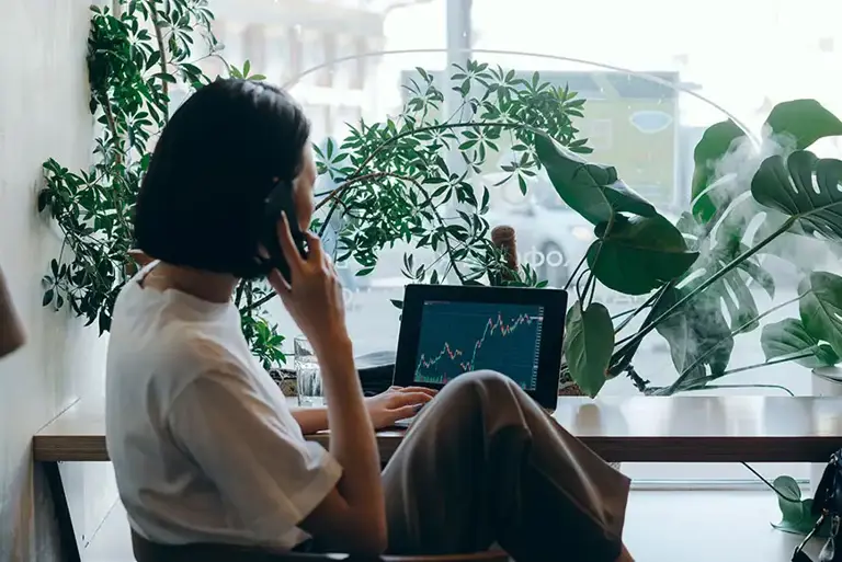 Woman seen from behind, sits at a table filled with green plants, works at a laptop showing charts 