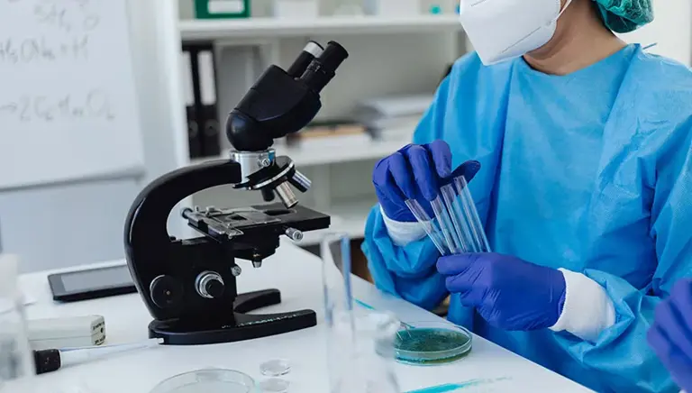 Masked scientist, in lab sat at a microscope holding a set of vials