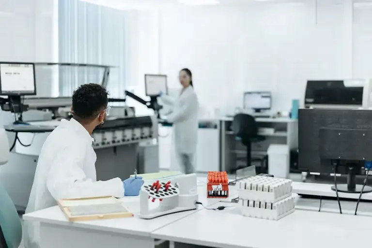 Two scientists in large lab, the one in the foreground is working on blood vials looks at the second in the background, while they work on at a large machine