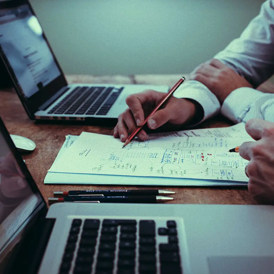 Close-up of the hands of two office workers writing notes on a desk between to open laptops