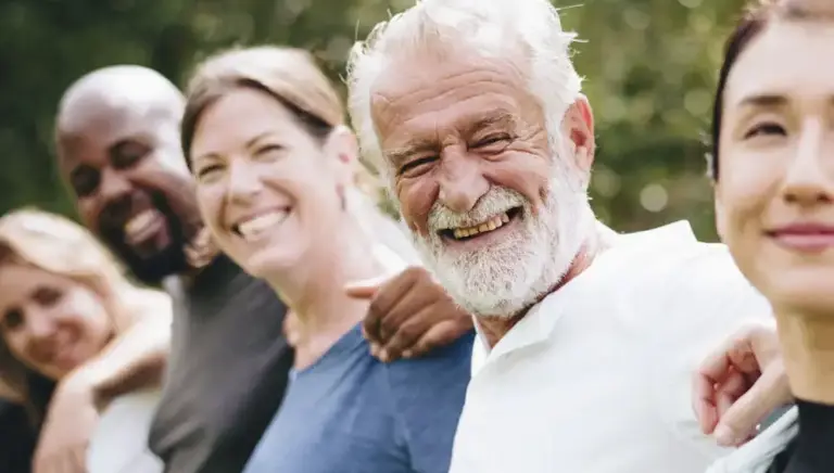 A line of people of all ages and diverse ethnic backgrounds smile to the camera while they all have their arms around each other's backs